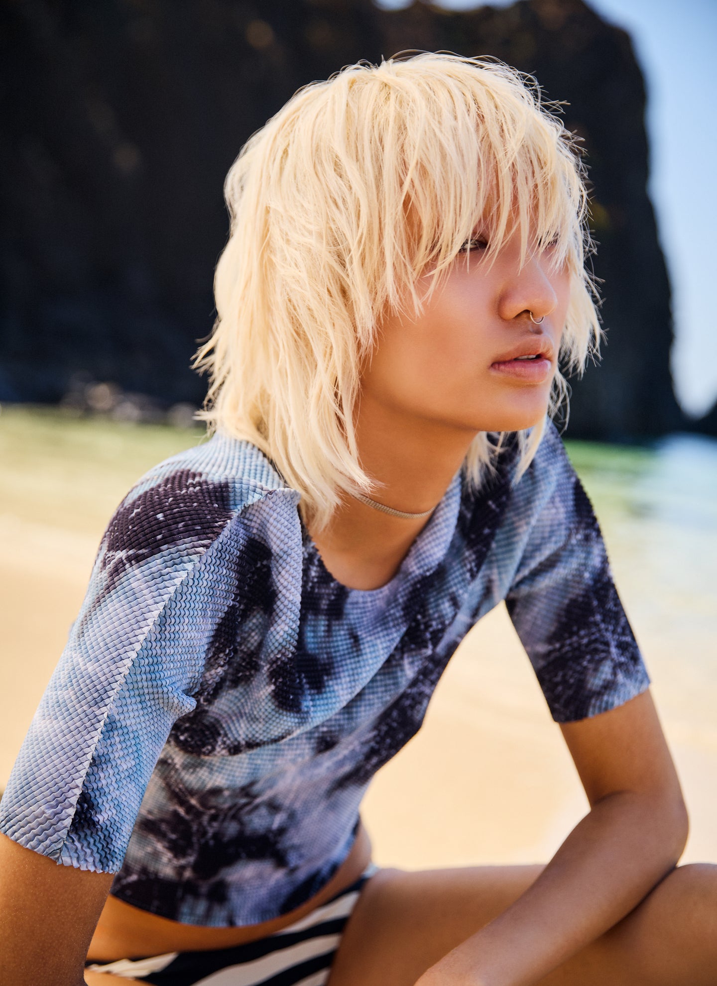 Person with blonde hair wearing a patterned shirt sitting on a beach.