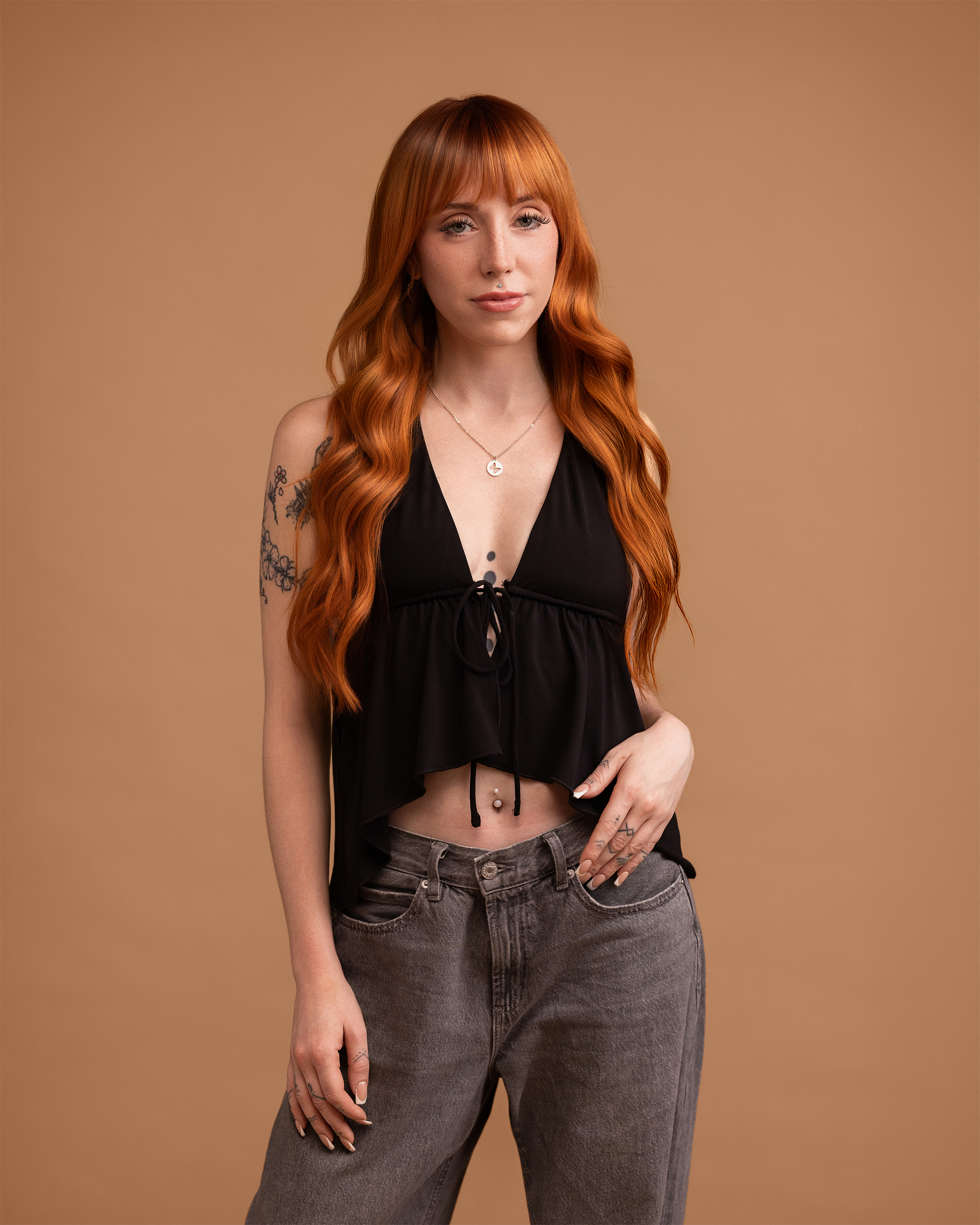 Woman with long red hair wearing a black top and gray jeans against a brown background