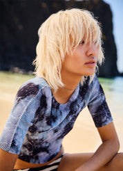 Person with blonde hair wearing a patterned shirt sitting on a beach.