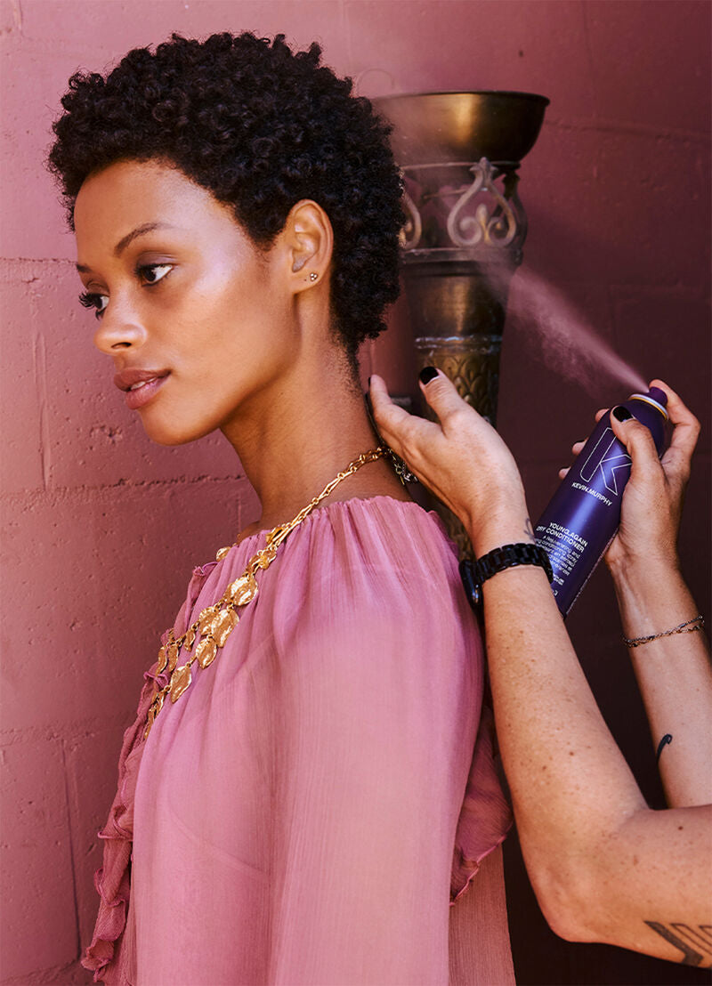 Woman applying hair product to her hair against a pink wall