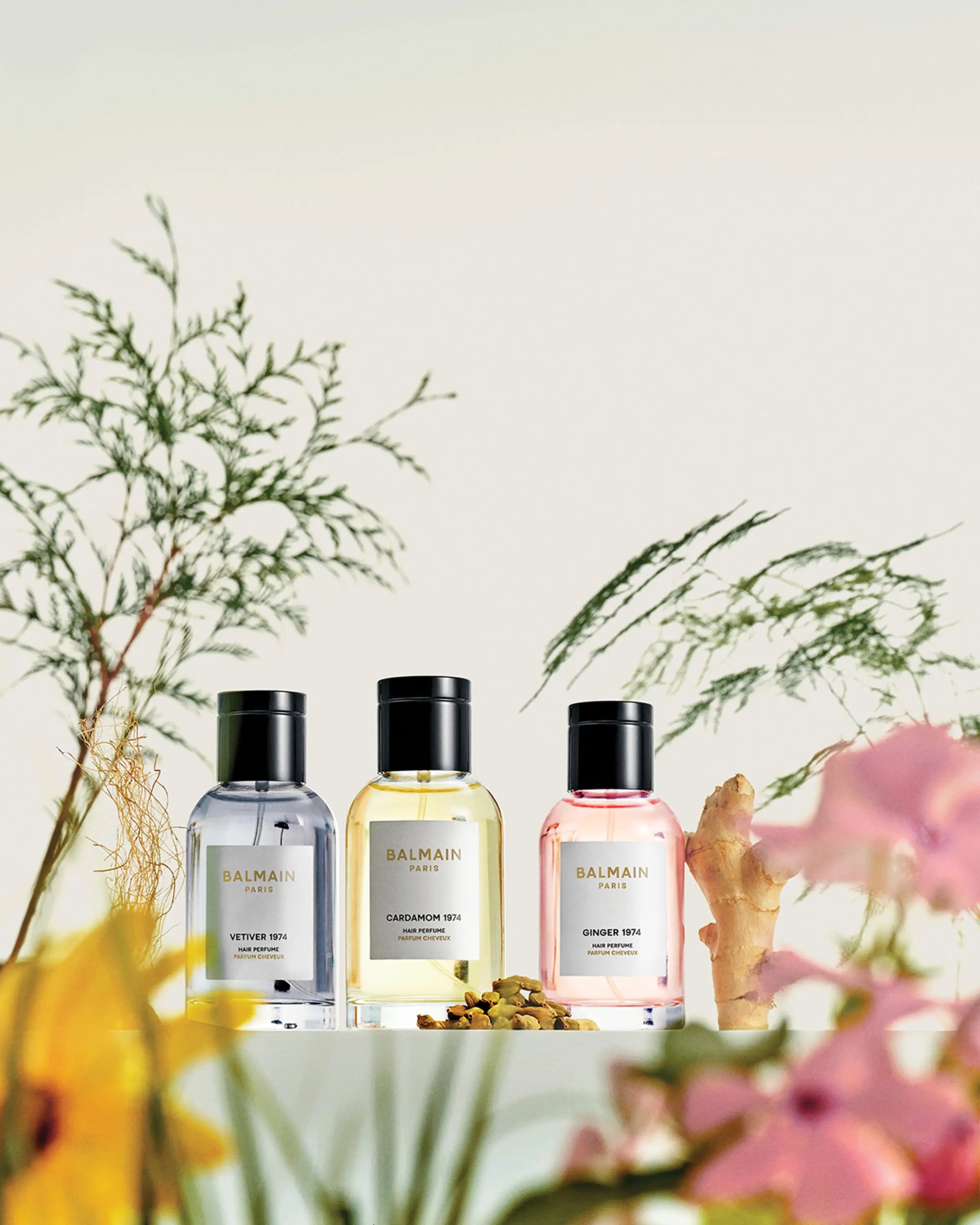 Three Balmain perfume bottles on a white surface with flowers and greenery in the background.